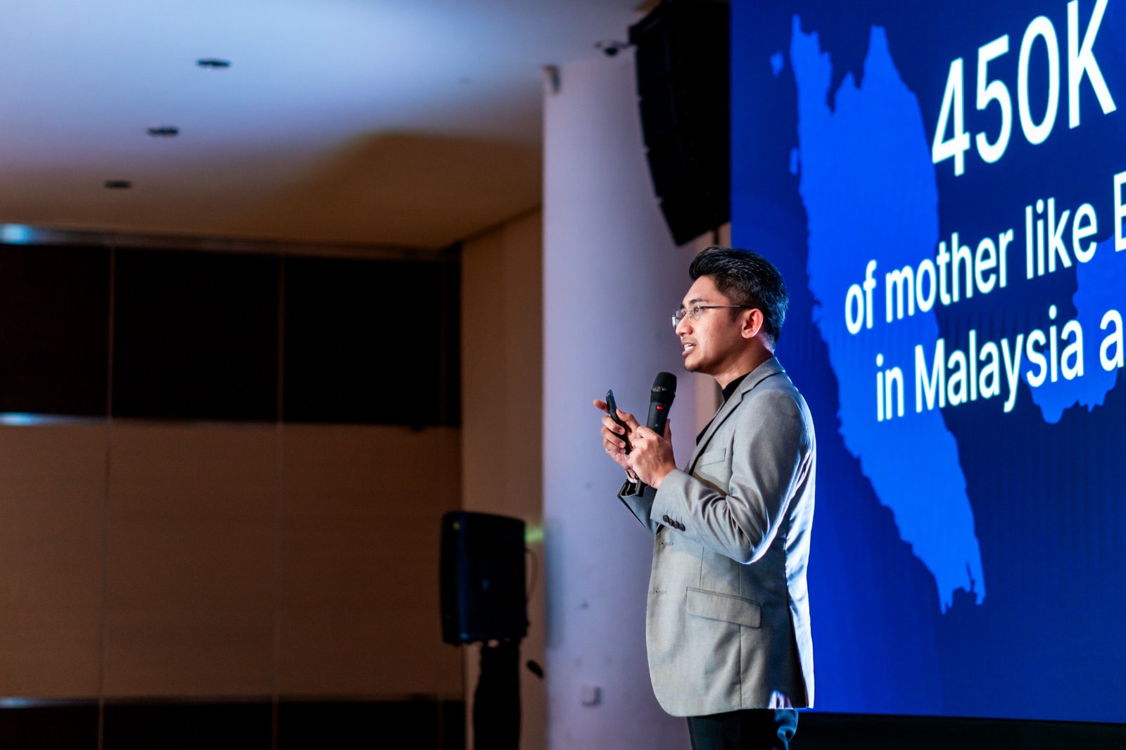 Shafeeq Zulkifli presenting on stage, wearing a grey suit and holding a microphone.